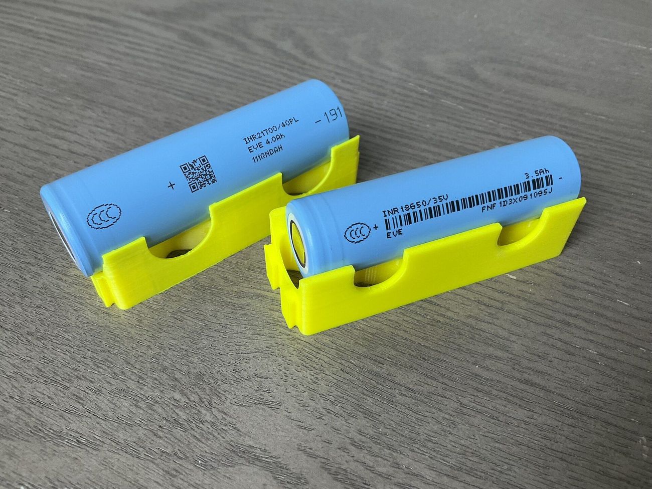 Two blue cylindrical lithium-ion cells, sizes 21700 and 18650, resting in yellow holders