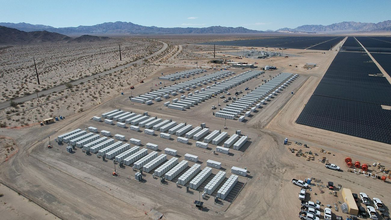 Aerial view of rows of white battery storage containers in the desert next to a large solar farm